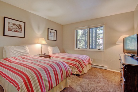 A bedroom with two single beds, each with red, gray, and white striped bedding. The room features a window, two bedside lamps, a dresser with a TV, and wall art above the beds.