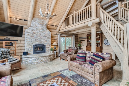 A spacious log cabin living room with exposed beams, a stone fireplace, mounted TV, and rustic furniture including patterned sofas and wooden decor. A staircase leads to an upper loft area.