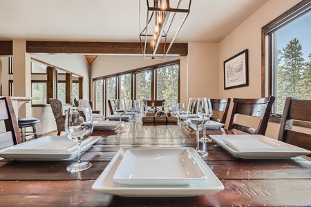 A wooden dining table set with square plates, wine glasses, and silverware in a modern dining room with large windows and a hanging light fixture.