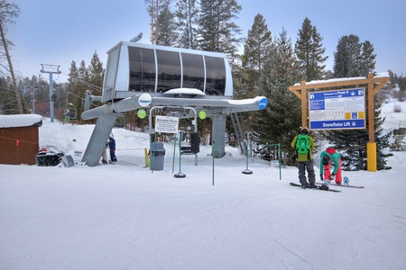 A ski lift station with people in ski gear preparing to board. There's a signboard on the right, and snow-covered trees in the background.