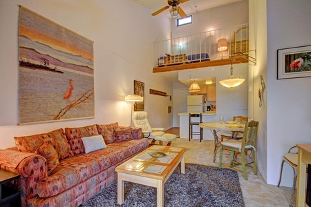 A cozy living room featuring a patterned sofa, glass coffee table, wall art, and an overhead loft with additional seating. The dining area, kitchen, and barstools are visible in the background.