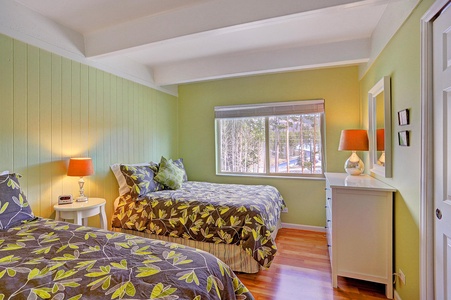 A bedroom with green walls features two single beds with leaf-patterned bedding, a bedside table with a lamp, a dresser with a mirror and lamp, and a window with a view of trees outside.