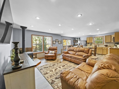 A cozy living room with beige leather sofas, an area rug, and a flat-screen TV, leads into an open, well-lit kitchen with light wood cabinetry and modern appliances.