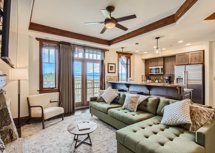 Modern living room with a green sectional sofa, round coffee table, and a wooden armchair, open to a kitchen area with a breakfast bar, stainless steel appliances, and large windows revealing an outdoor view.