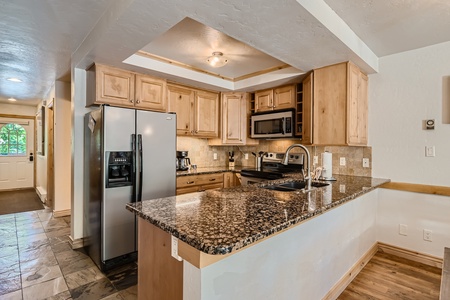 Modern kitchen with granite countertop, stainless steel appliances, wood cabinetry, tile backsplash, and a sink. The kitchen is open to a hallway and has a mix of tile and wood flooring.