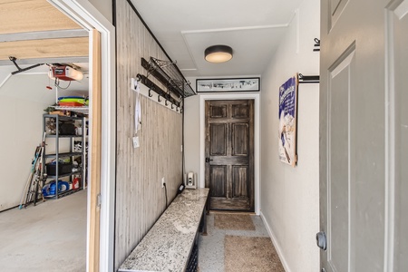 Entryway with a door leading to a garage, featuring a long bench, coat hooks, and storage shelves. A wooden door at the end of the hallway is closed, and various items are stored on the shelves in the garage.