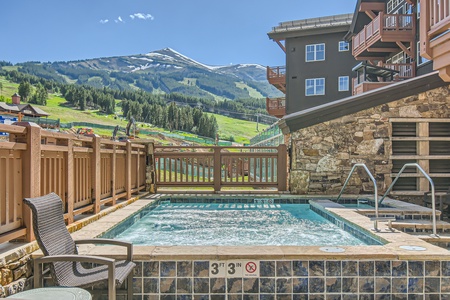 Outdoor hot tub with clear water, surrounded by a wooden fence with mountains and a resort building in the background. A sign indicates the water depth is 3 feet 3 inches.