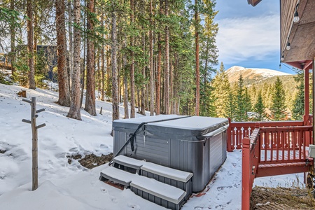 Outdoor hot tub with snow-covered steps, situated near a red wooden deck, surrounded by tall pine trees and mountains in the background.