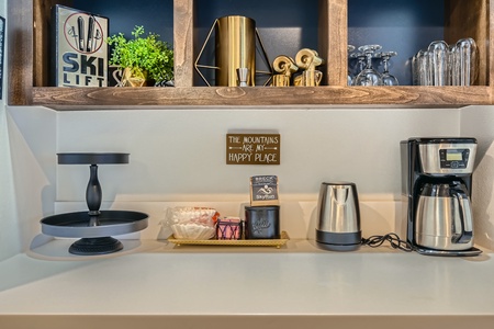 Kitchen counter with a coffee maker, kettle, tiered tray, and various decorations including a small plant, glasses, and a sign reading "The mountains are my happy place.