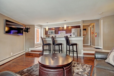 A modern living room featuring a mounted TV, a round coffee table, a sectional sofa, and an adjacent kitchen with a breakfast bar and three barstools.