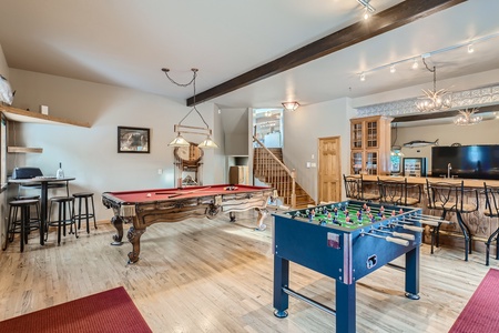 A spacious game room with a pool table, foosball table, bar area with stools, TV, and a staircase leading to another level. The room has wooden flooring and warm lighting.