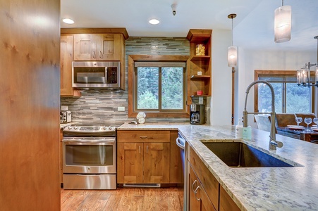 A modern kitchen with stainless steel appliances, wooden cabinets, a granite countertop, a window above the sink, and pendant lighting.