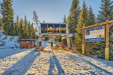 A ski lift station, labeled as "Snowflake Lift," is situated amidst snow and surrounded by trees. The station features a glass building and a nearby information board.