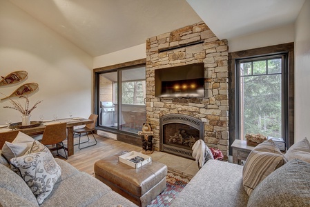 A cozy living room features a stone fireplace, wall-mounted TV, sectional sofa, dining area, and large windows overlooking a wooded area. Snowshoes hang on the wall, and a patio with a grill is visible.