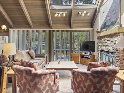 Living room with high wooden ceiling, large windows, sliding glass doors, fireplace, wall-mounted TV, sofa, armchairs, coffee table, and outdoor view of trees.