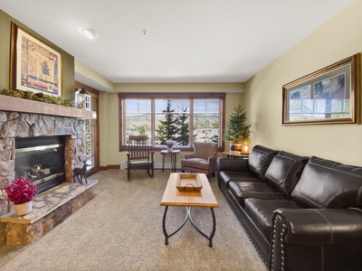 A cozy living room with a stone fireplace, black leather sofa, wooden coffee table, and large window offering a view of a tree. Decor includes framed artwork and a small Christmas tree.