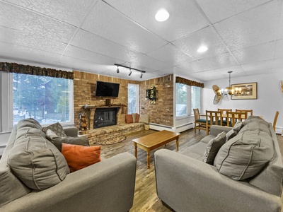 A living room with gray sofas, wooden coffee table, brick fireplace, wall-mounted TV, and large windows with white curtains next to a dining area with a wooden table and chairs.