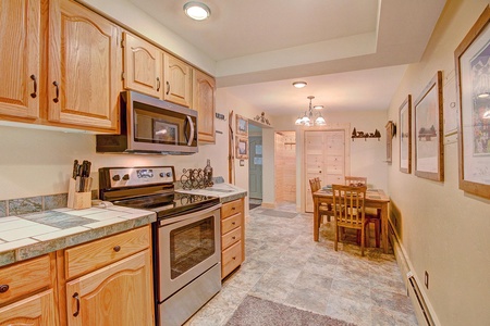 A kitchen with wooden cabinets, stainless steel stove and microwave, tiled countertops, and a dining area with a wooden table and chairs. Walls are decorated with framed pictures.