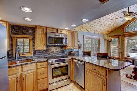 A modern kitchen with wooden cabinets, stainless steel appliances, granite countertops, a large island, and a vaulted ceiling with wood paneling. Skylights and large windows provide ample natural light.