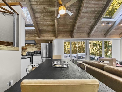 A modern, open-concept kitchen and living space with vaulted wooden ceilings, a ceiling fan, large windows, a black dining table, and a beige couch. Natural light illuminates the room.