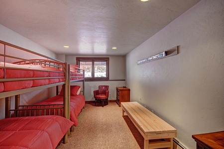 A room features two sets of red twin bunk beds on the left, a window with tall snow banks outside in the center, a small sitting area, a wooden bench, and a long coat rack on the right wall.