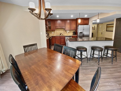 A kitchen with dark wood cabinetry, stainless steel appliances, and a breakfast bar with stools beside a dining table with chairs in a dining area with a chandelier.