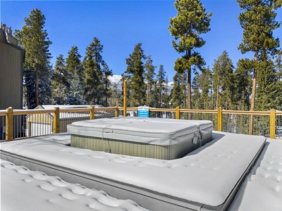 Snow-covered hot tub on a rooftop deck with a wooden railing, surrounded by tall trees and a clear blue sky in the background.