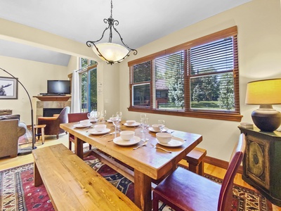 Dining area with a wooden table set for six, positioned near large windows with open blinds. A living area with a fireplace and TV is visible in the background.