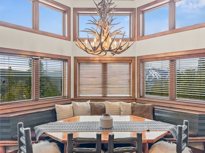 A cozy dining area with a wooden table, cushioned bench seating, and an antler chandelier. Large windows with wooden blinds offer a view of trees and sky.