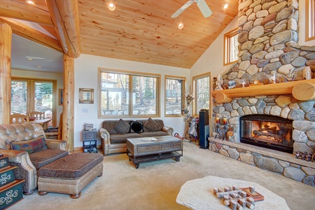 A cozy living room features a stone fireplace, wooden ceiling, and large windows. There is comfortable seating, including a sofa and an armchair with an ottoman, along with a central coffee table.