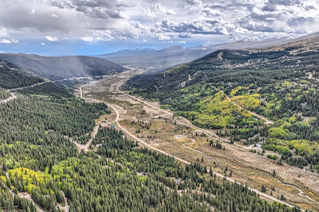 Aerial view of a mountainous landscape with winding roads, dense green forests, scattered yellow foliage, and a cloudy sky.