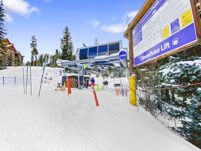 A snow-covered ski lift station named "Snowflake Lift" with people preparing to board. There are instructions and a trail map posted on a large sign on the right. Trees and buildings are in the background.