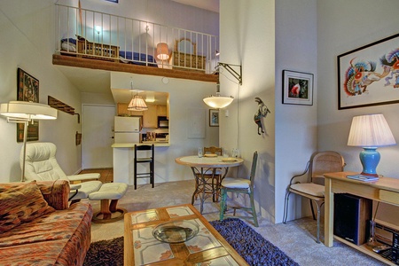 A cozy living space featuring a loft bedroom, a compact kitchen with white appliances, a dining area, a comfy sofa, armchairs, and wall art. The neutral color scheme and soft lighting create a warm ambiance.