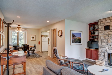 A living room with a stone fireplace, built-in bookshelf, and a TV. To the left, there is a dining area with a ceiling fan and a view into the kitchen. The room features wooden and cushioned furniture.