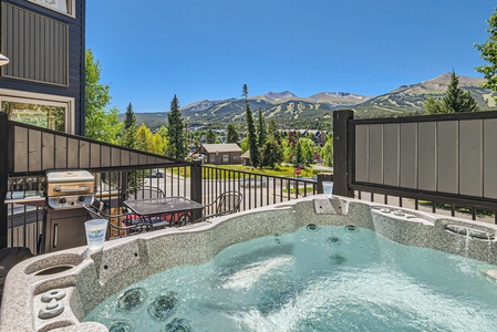 Outdoor balcony with a hot tub overlooking a mountainous landscape on a clear day. Grill and seating area are visible in the background.