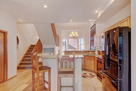 A kitchen and dining area with light wood cabinets, granite countertops, and a black refrigerator. A staircase leads to the upper floor, and a seating area with high chairs is in the foreground.