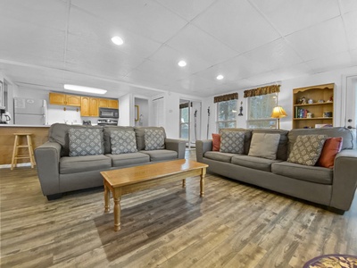 A living room with two gray sofas, a wooden coffee table, and a kitchen with wooden cabinets in the background. The room has wooden flooring and a bookshelf with decor items.