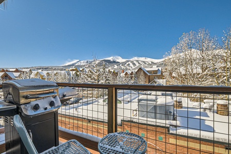 Snow-covered balcony with a grill and patio table set, overlooking a winter mountain landscape and buildings in the background.