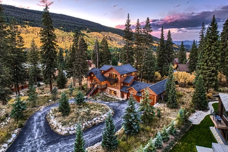 A large wooden cabin is surrounded by pine trees in a mountainous area at sunset. The driveway is curved and the sky has pink and purple hues.