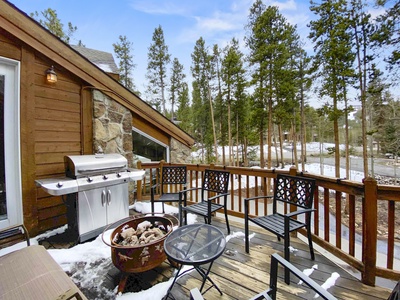 Wooden deck with a metal grill, fire pit, and seating area, surrounded by snowy trees. A house with a stone wall is on the left, and a forest view extends in the background.