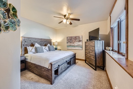 A cozy bedroom with a wood-framed bed, white bedding, bedside tables, a tall dresser with a TV on top, a ceiling fan, a window with blinds, and wall art decor.