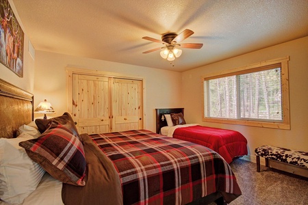 A bedroom with a double bed covered in plaid bedding and a single bed with red bedding, a wooden ceiling fan, and a large window with a forest view.