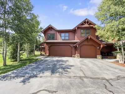 A two-story brown house with a double garage, surrounded by trees, on a sunny day.