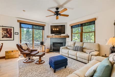 A cozy living room with two large windows, beige sofas, a blue ottoman, a leather armchair, a wooden chest, ceiling fan, and a wall-mounted TV above a fireplace. Trees are visible outside.