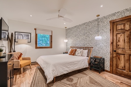 A bedroom with a white quilted bed, wooden headboard, patterned accent wall, wooden door, hanging pendant lights, and a window with a nature view. The room has light wood flooring and rug.