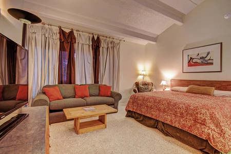 A bedroom with a large bed, couch, wooden coffee table, TV, standing lamp, wall art, two table lamps, and a large window with curtains.