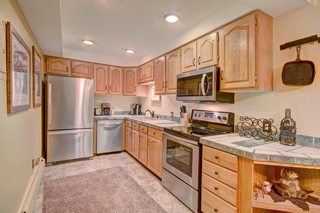 A modern kitchen with stainless steel appliances, wooden cabinets, a tiled countertop, and a carpeted floor. It includes a refrigerator, electric stove, microwave, and dishwasher.