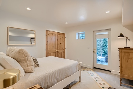 A cozy bedroom with a white bed, wooden dresser, and a small window. A door with glass panes opens to an outdoor view of trees. The room features recessed lighting and light wood decor.