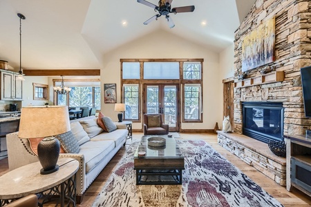 A living room with a high ceiling features a stone fireplace, large windows, and a ceiling fan. There is a white sofa, a wooden coffee table, a patterned rug, and wall art above the fireplace.