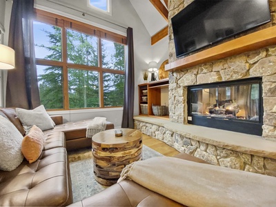 A cozy living room with a stone fireplace, large windows, a sectional sofa, and a wooden coffee table. A flat-screen TV is mounted above the fireplace.